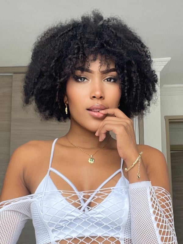 A woman with voluminous curly hair posing thoughtfully with her finger on her lips, wearing a white lace top and jewelry in an indoor setting.