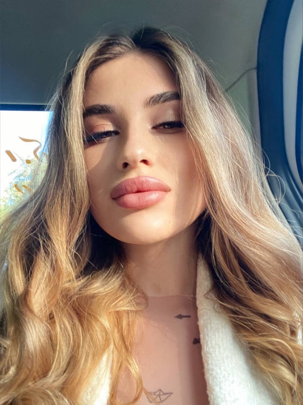A young woman with long blonde hair and full lips posing in a car, wearing a light-colored fuzzy top.