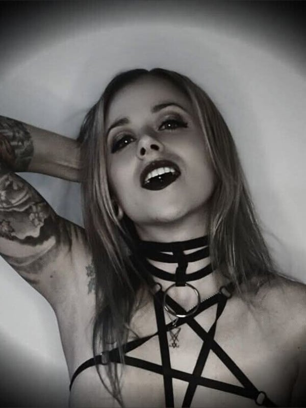 A woman with blonde hair, tattoos, and dark lipstick poses confidently in a black harness outfit with pentagram design, against a dark gradient background.