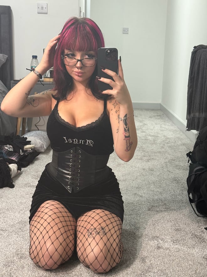 A person with magenta-streaked dark hair, glasses, facial piercings, and tattoos taking a mirror selfie indoors.