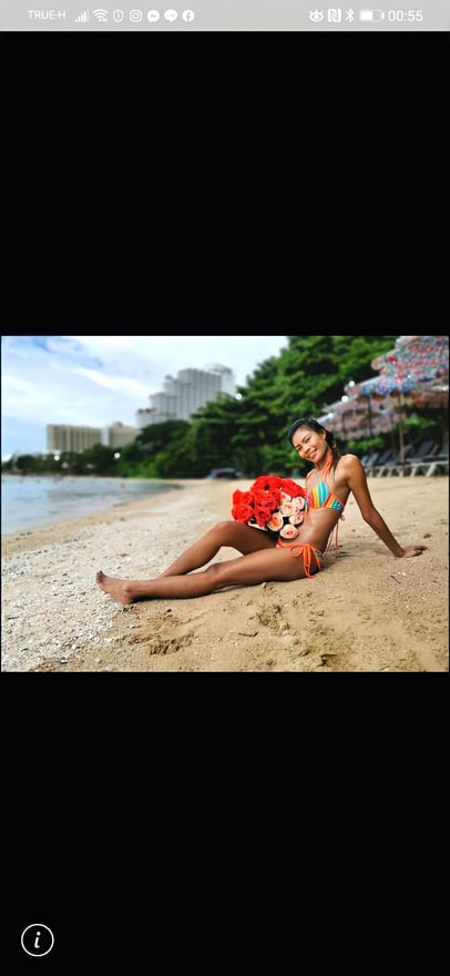 A woman in a colorful bikini sitting on a sandy beach, holding red flowers, with a scenic background of greenery and buildings.