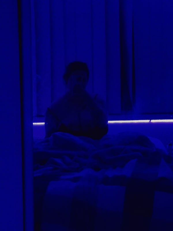 A silhouette of a person sitting on a bed with purple lighting and blinds.