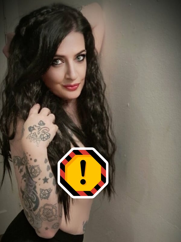 A woman with long dark wavy hair and tattoos on her arm, posing confidently with a warning sign overlay.