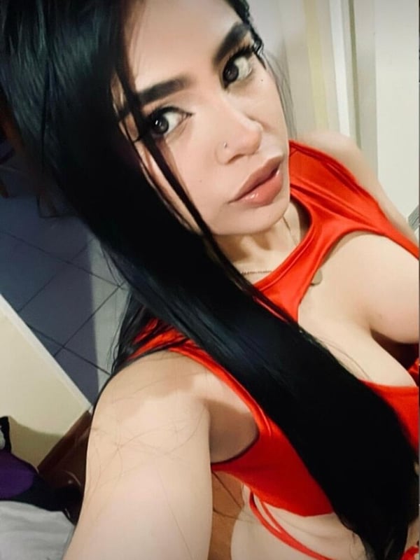 Young woman with long dark hair taking a selfie in a room, wearing a red top, posing confidently.