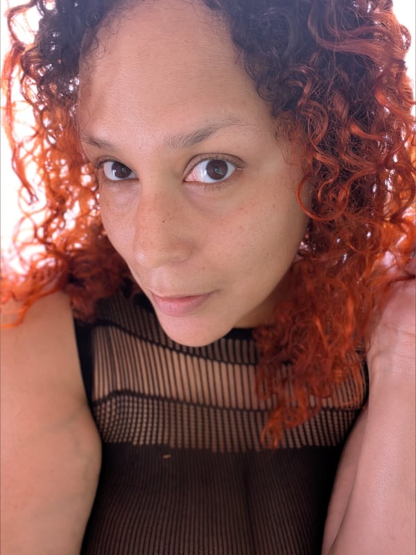 Woman with curly reddish hair smiling at the camera, wearing a black mesh top against a white background.