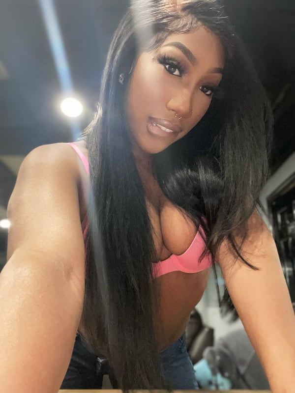 A woman with long dark hair takes a close-up selfie in a pink top, smiling at the camera in an indoor setting with soft lighting.