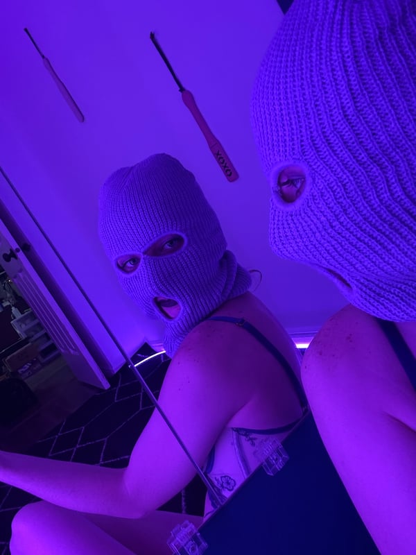 Two people wearing balaclava masks in a purple-lit room with neon accents, sitting close together.
