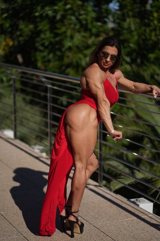 A muscular woman in a red dress poses confidently on a balcony overlooking lush green trees and a railing.