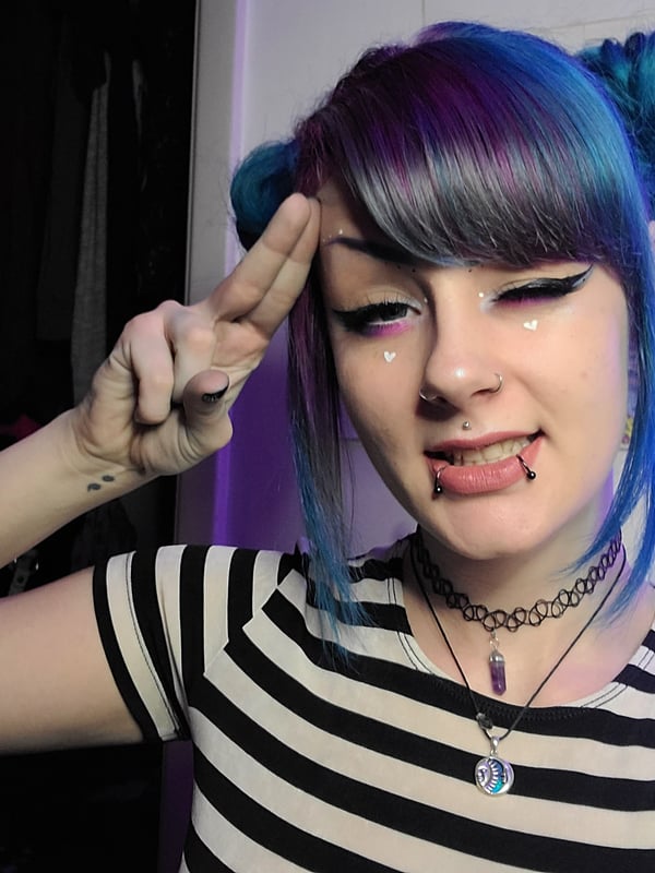 Young woman with colorful dyed hair in pigtails making a playful gesture with tongue out, wearing a striped shirt and choker necklace indoors.