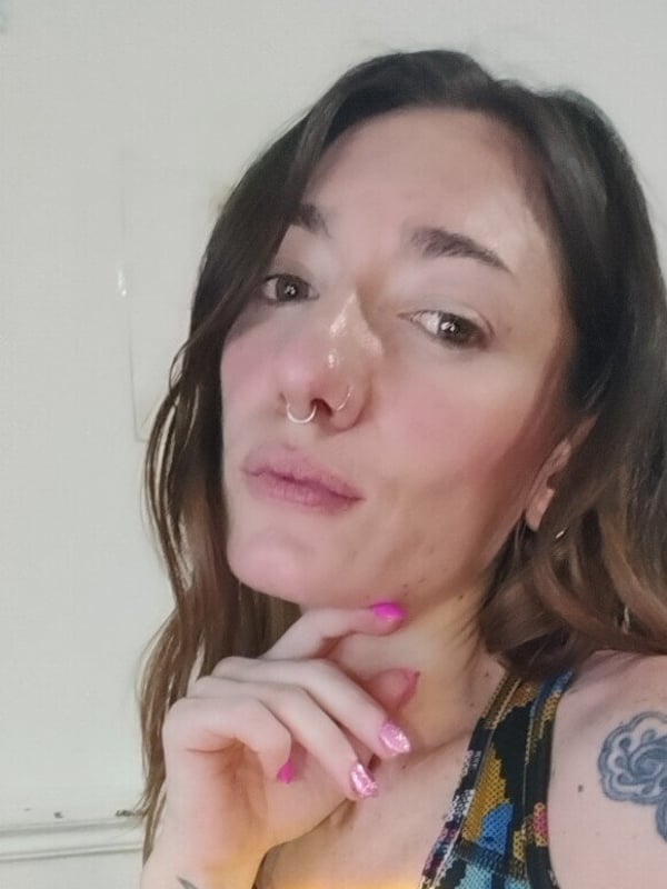 A woman with freckles, tattoos, and pink nails posing thoughtfully in a colorful top against a plain background.
