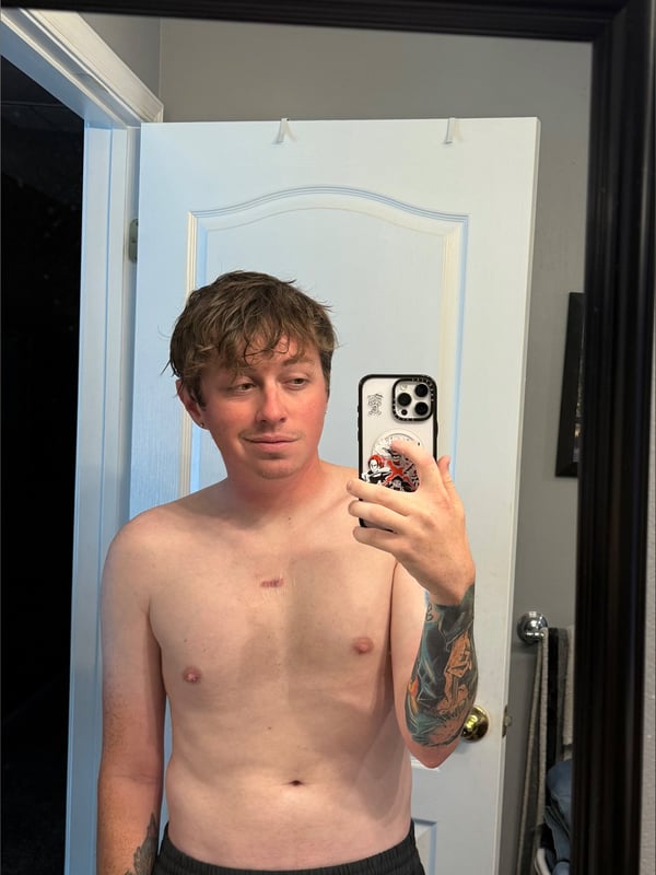 A person taking a shirtless selfie in a bathroom mirror, showing tattoos on the arm, wearing sweatpants.