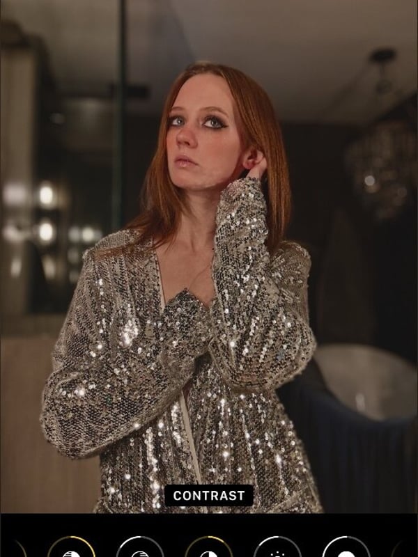 Close-up of a woman with red hair and subtle makeup, wearing a shimmering top, captured in a mirror reflection with photo editing tools visible.