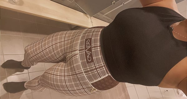 A woman standing in front of a mirror, wearing a black tank top and plaid pants with a belt.