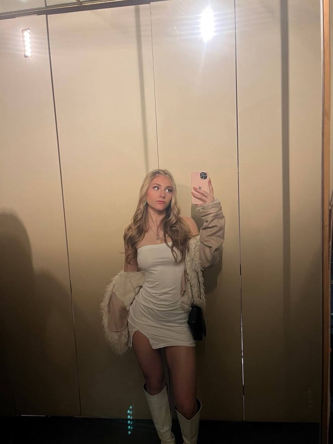 A young woman taking a selfie in a white satin crop top with a small tie detail, against a plain wall.