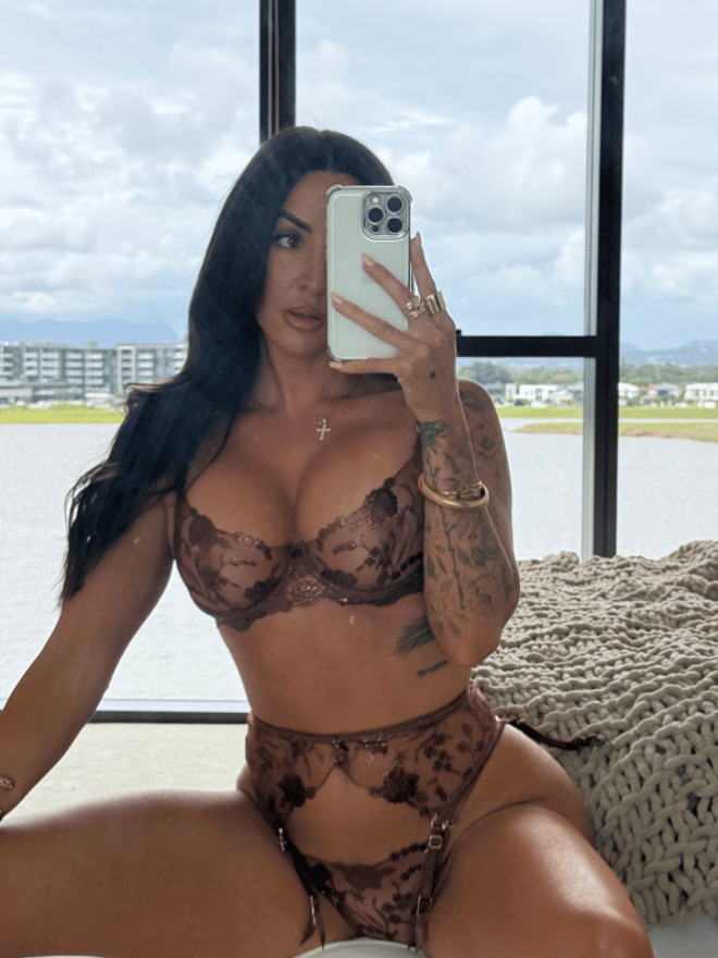 A woman with long dark hair posing in a bathroom wearing red and black lingerie, showing tattoos on her back and arms.