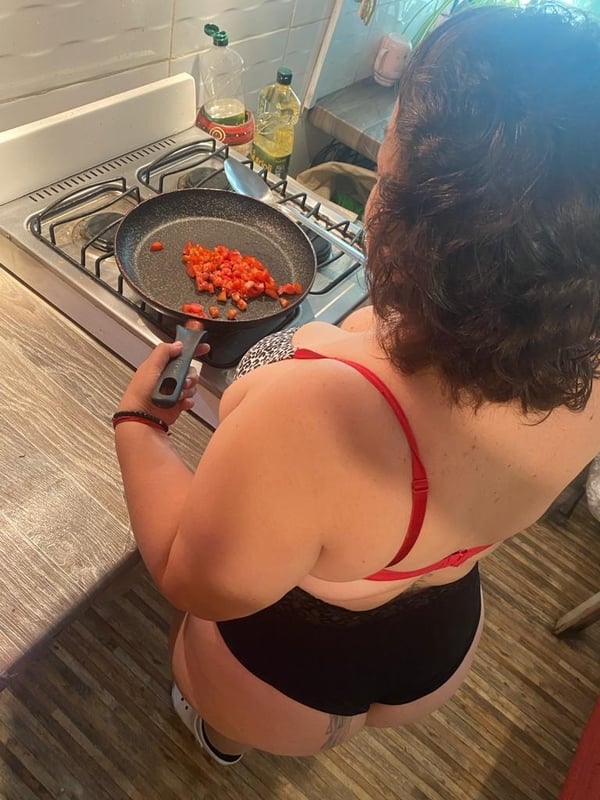 A woman with curly hair cooking peppers on a stovetop in a kitchen, wearing a red top and black bottoms.