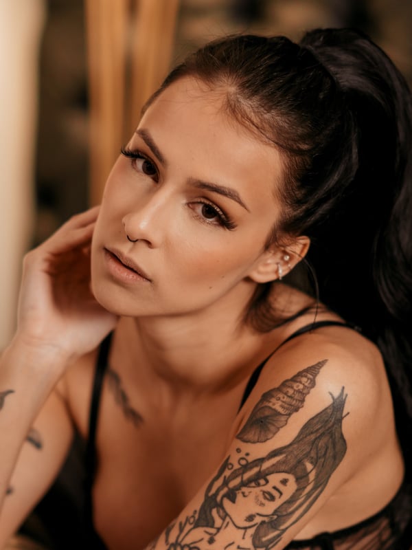 Close-up portrait of a woman with long dark hair in a ponytail, wearing lace top, with tattoos on her arm and shoulder, posing thoughtfully indoors with warm lighting.