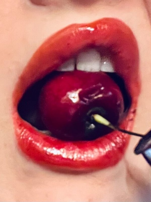 Close-up of a woman's face with glossy red lips holding a cherry in her mouth using a toothpick.