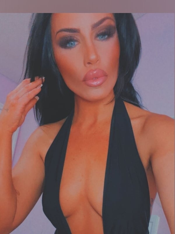 Woman with long dark hair and bold makeup posing in a black halter top against a purple background.