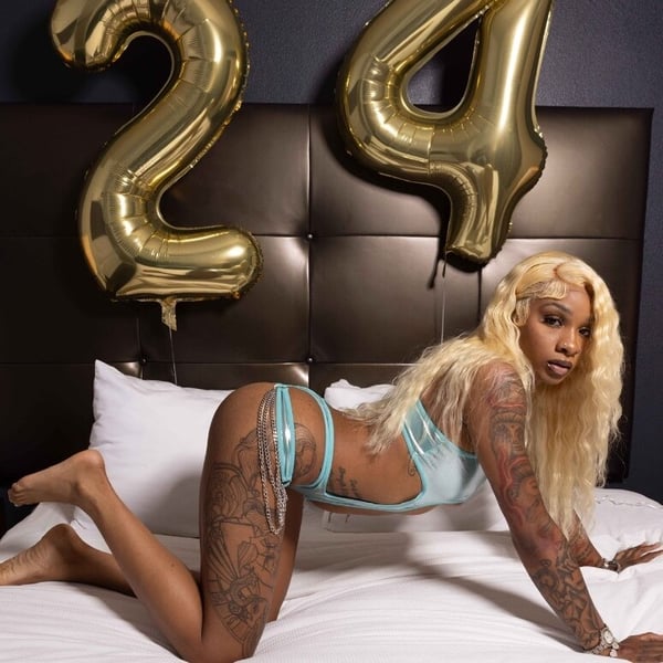 A woman with long blonde hair posing on a bed surrounded by large gold '24' balloons against a dark headboard.