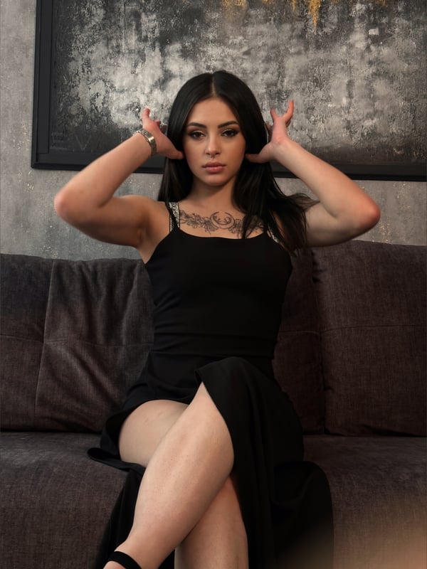 Young woman with long dark hair sitting on a couch, wearing a black spaghetti strap top and skirt, making hand gestures in front of a textured wall art.