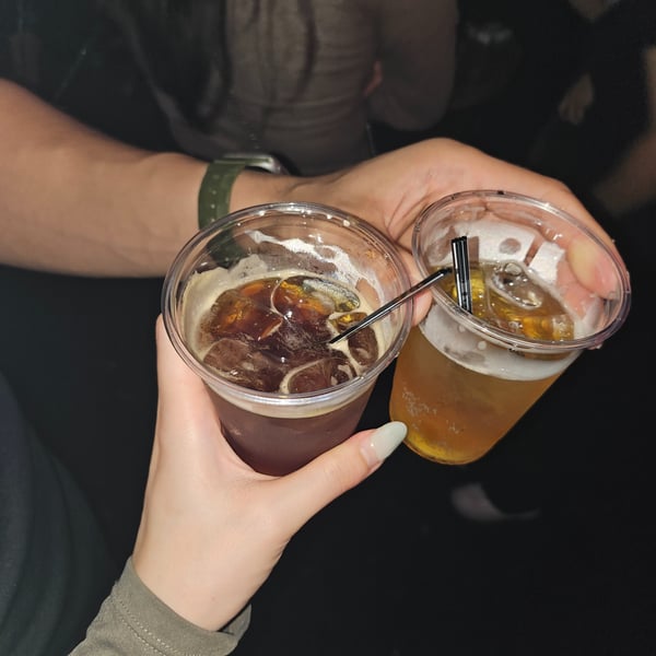 A couple holding plastic cups of iced coffee and beer in a dimly lit setting, toasting with smiles.