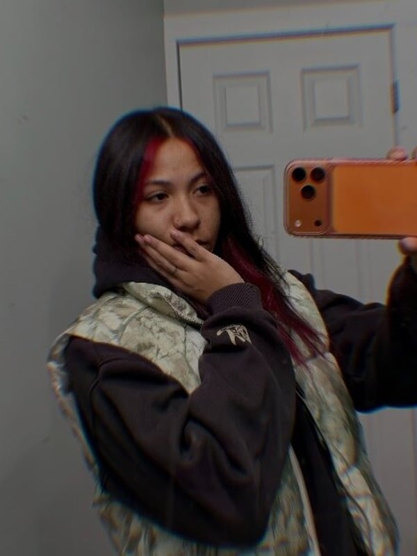 Young woman with red-streaked hair taking a surprised selfie in a hoodie while holding a phone.
