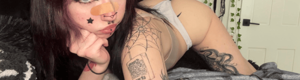 A person with purple hair and tattoos posing indoors on a bed.
