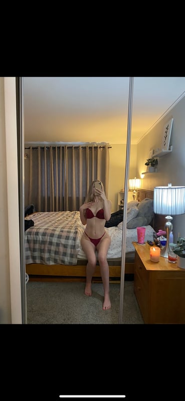 Young woman sitting on a bed in a red bikini, posing in front of a mirror in a cozy bedroom with warm lighting and curtains.