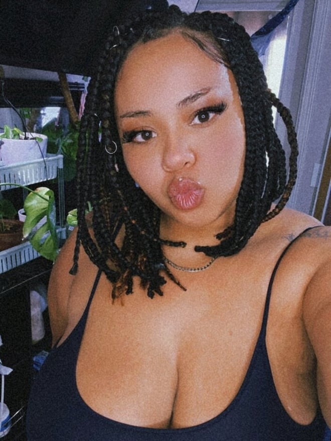 A young woman with braided hair and makeup, puckering her lips for the camera, standing in a room with plants and shelves.