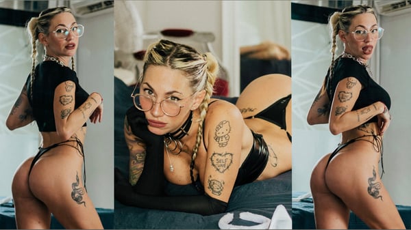 A woman with blonde hair and glasses posing confidently in stylish black outfits on a bed, showcasing tattoos and edgy accessories.