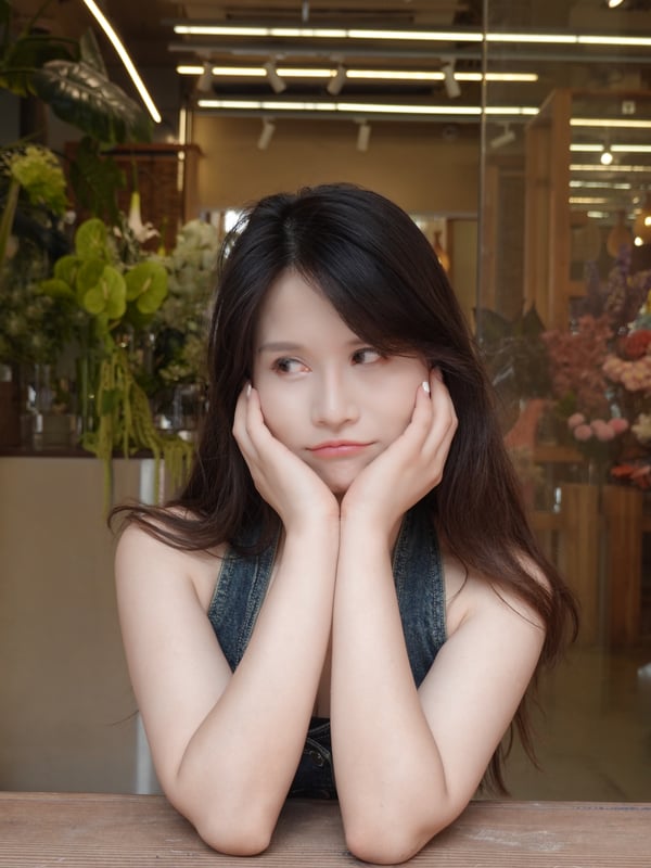 A woman with long dark hair wearing a light blue cardigan, leaning against a window frame indoors.