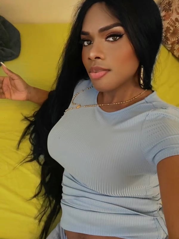 A woman with long black hair smiling at the camera, wearing a light blue top and shorts while lounging on yellow cushions.