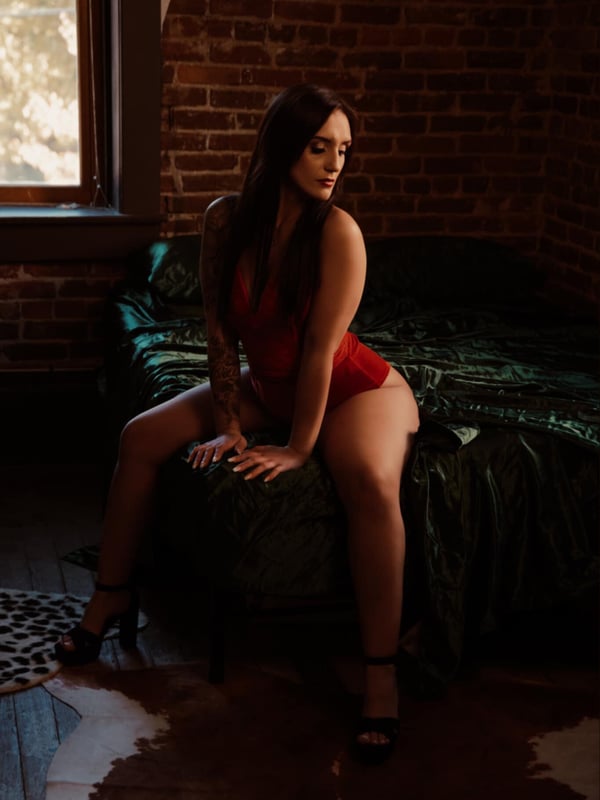 A woman with long dark hair sitting on a bed in a red outfit and high heels, against a brick wall with warm lighting.