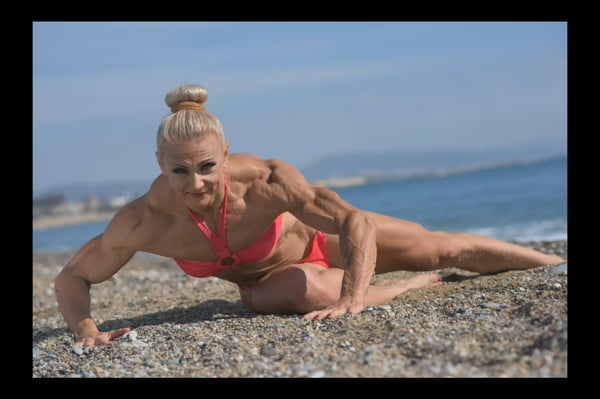 Muscular woman in a red bikini posing dynamically on a beach with ocean waves in the background.