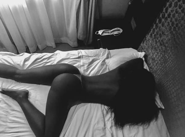 A person lying nude on a bed in a dimly lit room with sheer curtains and wooden furniture.