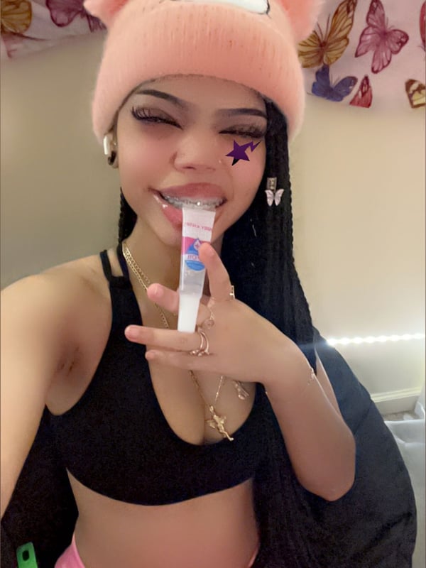 Young woman with long braided hair wearing a pink Hello Kitty beanie, black top, and pink bottoms, holding a tube while smiling in a room with pink decor.