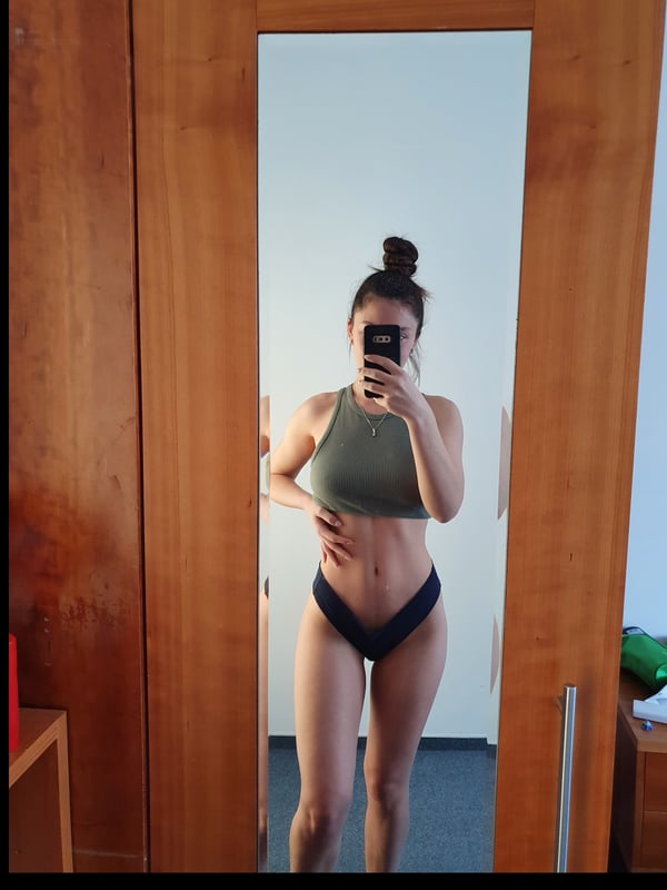 Young woman taking a selfie in front of a wooden wardrobe mirror, wearing a green tank top and black bottoms, showing her toned midriff.