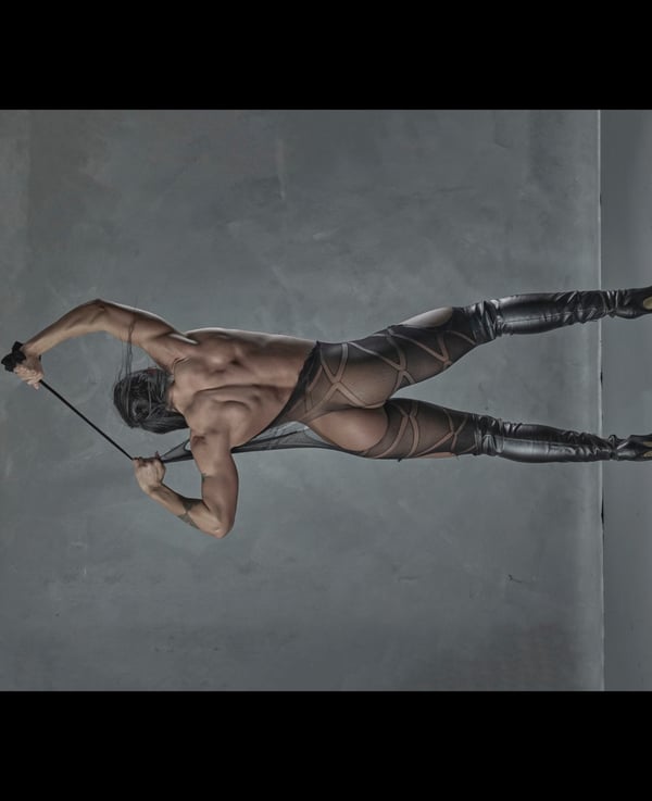 A muscular person in a dynamic pose wearing black latex attire and boots against a gray background.