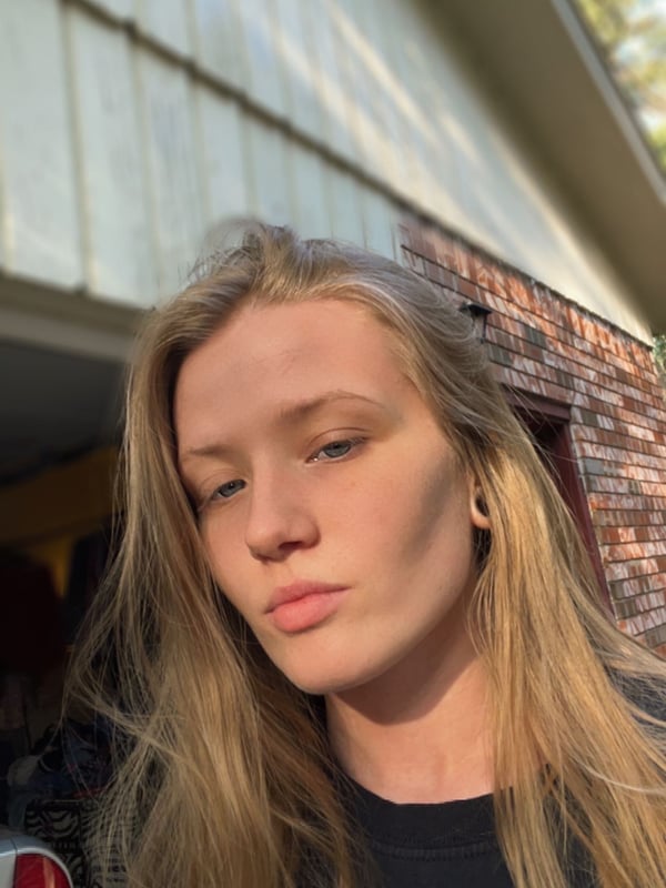 A young woman with long blonde hair standing in front of a garage door, wearing a black top, with a casual expression.