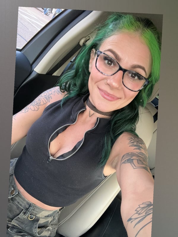 A woman with vibrant green hair and glasses smiles in a car selfie, wearing a black top and choker with visible tattoos.