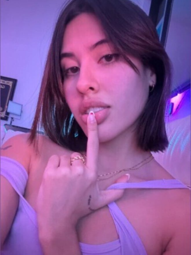 Young woman with short brown hair making a playful gesture with her finger to her lips, wearing a purple off-shoulder top in a room with neon lighting.