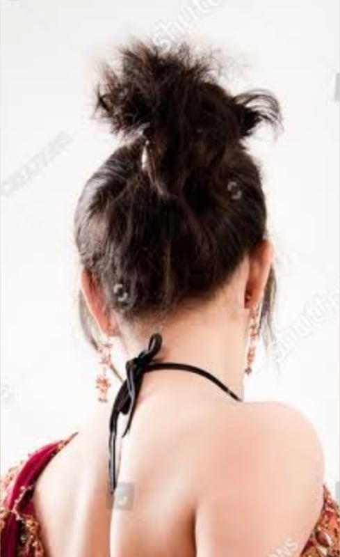 Woman with long dark hair in a messy bun, wearing a traditional embroidered outfit, posing with her back to the camera against a plain background.
