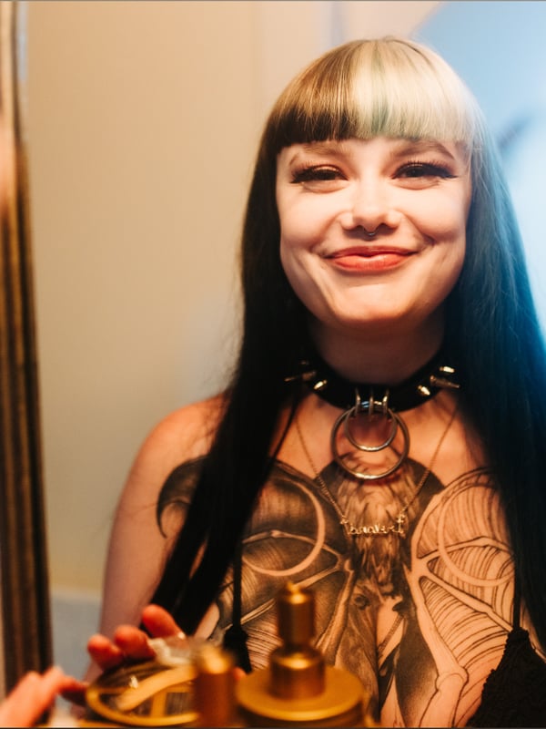 A woman with long dark hair, tattoos, and a choker necklace smiling in a mirror selfie inside a room with a photographer nearby.