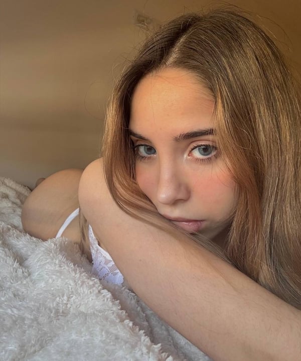Young woman with freckles and long wavy hair lying on a bed, posing sideways in white lingerie with a thoughtful expression.