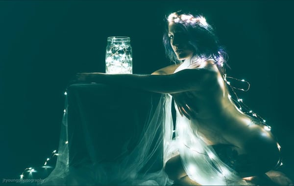A woman with flowing hair poses gracefully near a glowing jar on a draped table, surrounded by soft lights and fabric in a dark artistic setting.