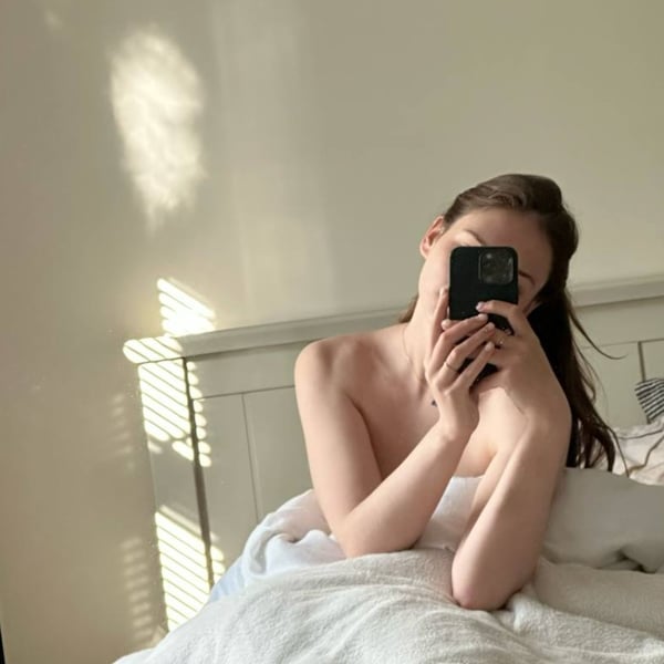 A young woman taking a selfie in a sunlit bedroom, sitting on a bed with white sheets and wooden headboard.