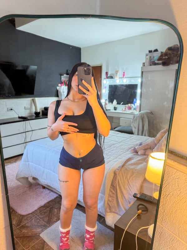A woman taking a mirror selfie in a modern indoor space, wearing dark underwear and showing visible tattoos on her arms and torso.