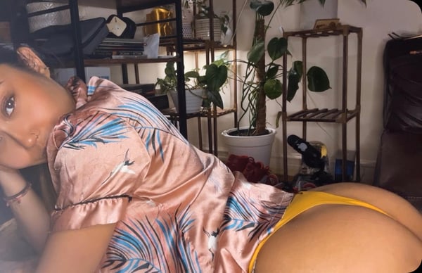A woman lounging on a bed in a silky floral robe and yellow bottoms, surrounded by plants and decorative items in a cozy room.