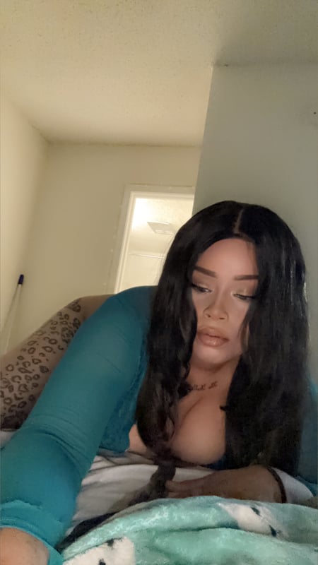 A woman with long dark hair posing on a bed in a sheer turquoise top inside a bedroom with neutral walls.
